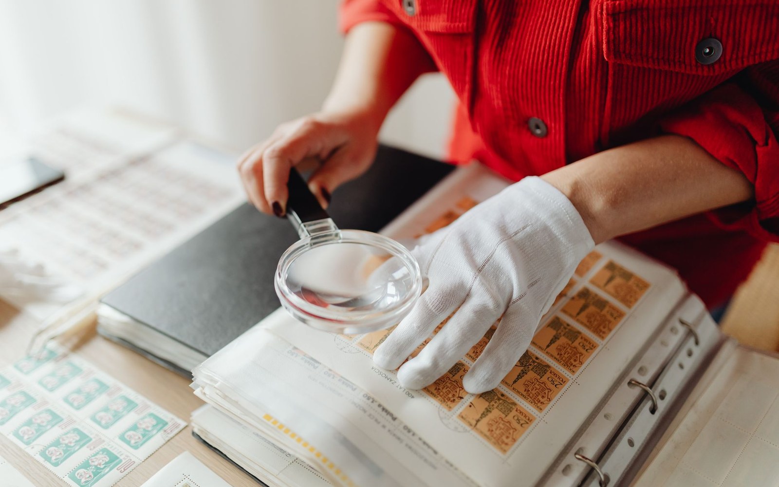 A gloved hand uses a magnifying glass to examine stamps in an album
