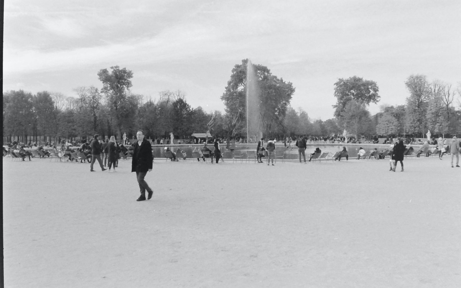 People gather around a fountain pool in a tree-lined park as a man walks past