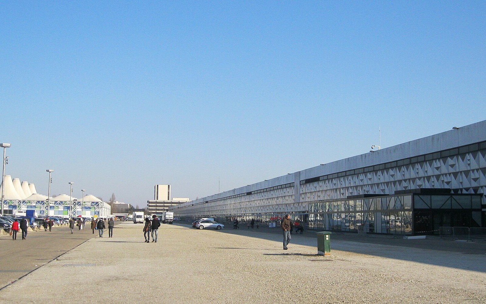 People walk along a gravel path beside a long low-rise exhibition building with a patterned facade