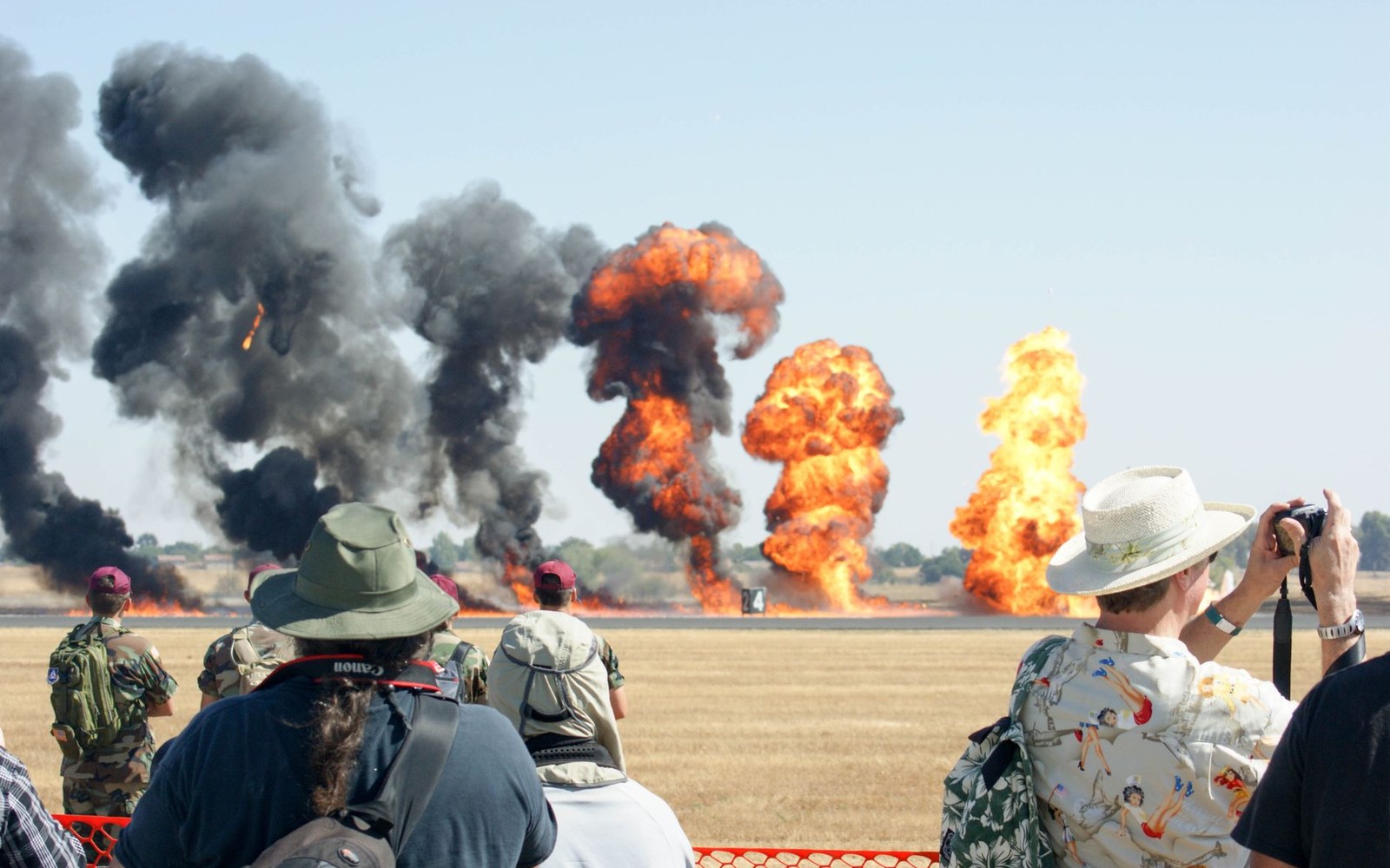 Spectators packed along the barrier at an air display, cameras up as the pyrotechnics go off down the runway.